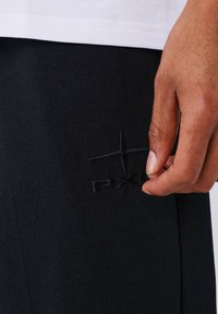 Pantaloni in tessuto nero con trama testurizzata; presentano un logo ricamato nero prominente e una mano che afferra il tessuto vicino al logo.