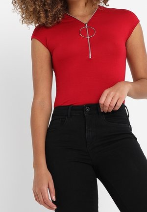 Frau trägt ein eng anliegendes rotes Oberteil mit einem silbernen Reißverschluss in Ringform und hoch taillierte schwarze Hose, posiert mit einer Hand in der Nähe der Taille.