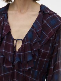 Persona, kas valkā bordo un zilu pleda blūzi ar volānu apkakli un mazu kaklasaiti dziļā V izgriezumā, redzama no pleciem līdz augšdaļai krūtīs.