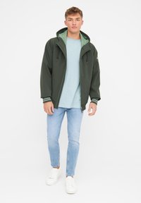 Dunkelgrüne Softshelljacke mit Kapuze, ausgestattet mit kontrastierender grüner Innenausstattung und elastischen Bündchen, kombiniert mit einem hellblauen Hemd und hellen Jeans.