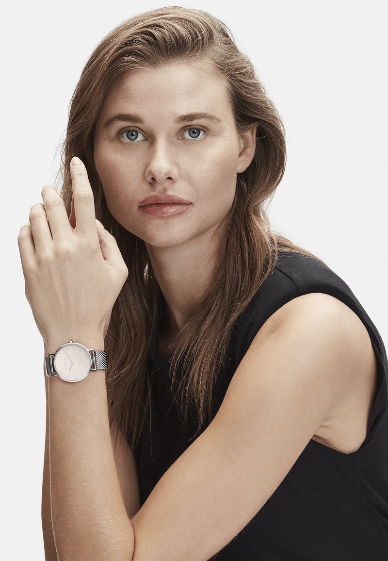 Zilveren gaas armbandhorloge met een ronde, lichtroze wijzerplaat, minimalistische uuraanduiders en een slank, reflecterend kastje.