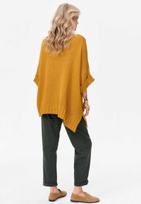 Musztardowożółte dzianinowe ponczo o luźnym kroju, z prążkowanym dołem i bocznymi rozcięciami, zestawione z ciemnozielonymi spodniami i jasnobrązowymi butami wsuwanymi.
