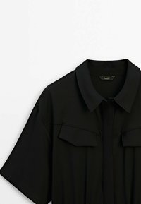 Zwart shirt met korte mouwen, een met kraag en twee borstzakken. Soepele textuur met een knoopsluiting aan de voorkant.