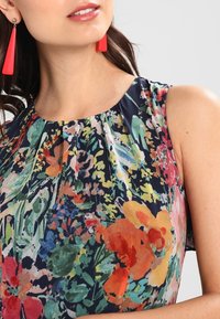 Kvinna i ärmlös blommig klänning med flerfärgade blommor och långa röda triangulära örhängen mot en ljus bakgrund.