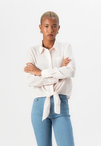 Lys rosa, langermet skjorte med knappestolpe og knyttet midje, med en brystlomme og subtil teksturert stoff. Kombinert med lyseblå jeans.