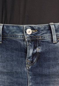 Gros plan de la taille d'un jean en denim bleu avec un bouton en métal et des passants de ceinture, porté avec une chemise noire rentrée.