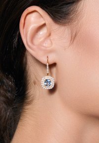 Złota kolczyka w kształcie kółka z dużym niebieskim kamieniem szlachetnym, otoczonym halo mniejszych, przezroczystych kamieni, o gładkim, wypolerowanym wykończeniu.