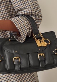 Melna ādas soma ar zelta detaļām, rakstura dizains, divi rokturi un dekoratīvi kuloni. Daļēji redzama rakstaina krekls.