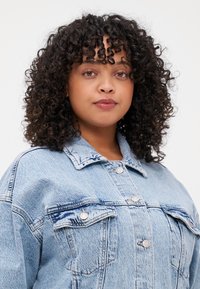 Chaqueta de mezclilla azul claro con un ajuste ligeramente holgado, que presenta dos bolsillos frontales, botones plateados y un diseño de cuello clásico.