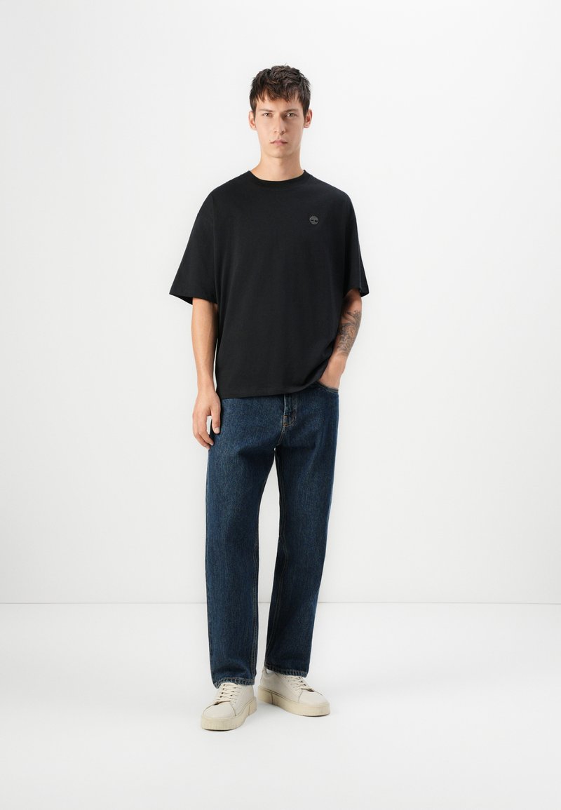 Czarna koszulka z krótkim rękawem i okrągłym dekoltem, teksturowana tkanina, w połączeniu z luźnymi niebieskimi dżinsami i jasnymi trampkami.