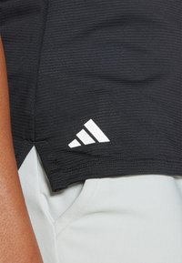 Črna športna majica z ribasto strukturo, ki ima belo Adidasovo logo na robu in je v kombinaciji z svetlo sivo tkanino spodaj.