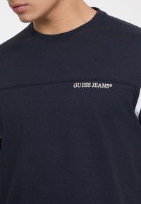 Tamsiai mėlynas medvilninis džemperis su paprastu apvaliu kaklu, turi baltą akcentinę juostą ir išsiuvinėtą „GUESS JEANS“ logotipą per krūtinę.