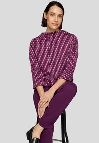 Fuchsia und weißes gemustertes Oberteil mit Boots-Ausschnitt und 3/4-Ärmeln; kombiniert mit lila Hosen und schwarzen Schuhen. Model sitzt auf einem Hocker.