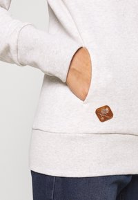 Jasnoszara bluza z ribbingowanymi mankietami i dołem. Posiada kieszeń z przodu oraz skórzaną metkę z logo z boku. Miękka w dotyku.