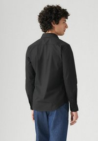 Camisa negra de manga larga con cuello abotonado, textura suave y dobladillo recto. El modelo lleva puesta una jeans azules.