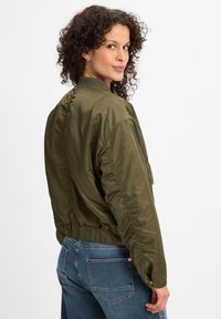 Maslinasto zeleni bomber jakna s glatkom, sjajnom teksturom, rebrastim ovratnikom, elastičnom trakom u struku i bočnim dzepovima. Model prikazan s leđa.