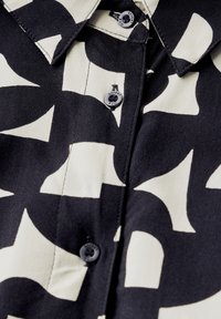 Camicia button-up nera e crema con un motivo astratto, tessuto con una texture liscia e colletto appuntito con due bottoni neri.