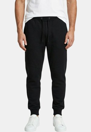 Homme portant un pantalon de jogging noir avec taille à cordon, poignets côtelés et baskets blanches, debout devant un fond uni.