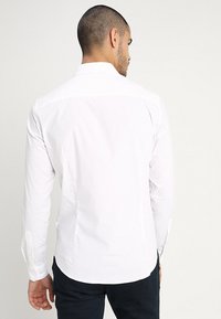 White long-sleeve shirt with a collar, smooth texture, and buttoned cuffs. Back view shows a tailored fit and gentle curved hemline.