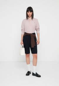 Chemise boutonnière rose rayée, shorts foncés, pull marron noué à la taille, sac à main blanc, mocassins noirs, chaussettes blanches. Style minimaliste, design épuré.