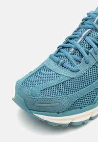Turkizni športni čevelj iz mrežastih in semiš materialov, z teksturirano kapico za prste, kontrastnimi črtami in vezalnim dizajnom.