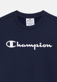 T-shirt blu navy in cotone con scollatura a girocollo, caratterizzata da un grande logo bianco "Champion" stampato sul petto in carattere corsivo.