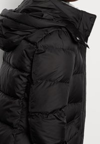 Zwarte pufferjas met capuchon, voorzien van een doorgestikte ontwerp, glanzende textuur en drukknopen. Zijkant toont dikke isolatie.