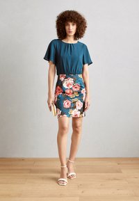 Tonga Bluse mit kurzen, schwingenden Ärmeln, kombiniert mit einem taillierten, türkisfarbenen Blütenrock mit rosa und gelben Rosen. Das Model hält eine kleine cremefarbene Clutch.