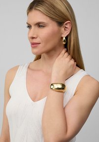 Gouden druppeloorbellen en een gladde gouden koppeltas gedragen door een model met lang steil haar, tegen een neutrale achtergrond.