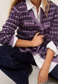 Žena s fialovým kardiganem s geometrickým vzorem přes bílou košili, v tmavě modrých džínách, sedí s jednou rukou položenou na kardiganu.