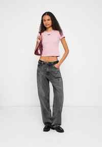 Šviesiai rožinė, glaudžiai prigludusi trumpa palaidinė su logotipo detalėmis, derinama su laisvais, blukintais juodais džinsais ir juodais slip-on batais. Rankose laikoma bordinė rankinė.