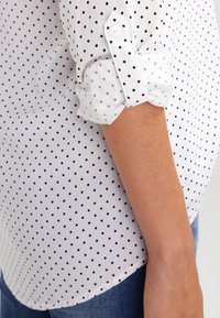 Camicia bianca con pois neri, dotata di maniche arrotolate, chiusura a bottoni e una texture liscia con un orlo curvo.
