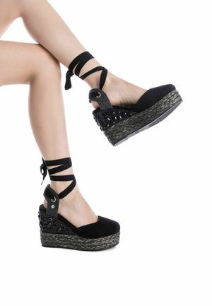 Sandalias de cuña negras con un upper de tela texturizada, tiras en los tobillos y una plataforma de cuerda trenzada. La suela presenta un diseño estampado.