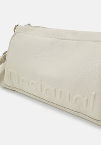 Cremefarbene Lederhandtasche mit geprägtem "Desigual"-Logo, glatte Textur, silberner Reißverschluss und rechteckige Form. Mit Akzentnähten entlang der Kanten.