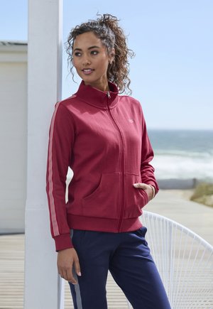 Red zippered jacket with a high collar, featuring two front pockets and white striped accents along the sleeves. Paired with navy pants.