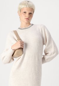 Šviesiai smėlio spalvos megztinis su pūstomis rankovėmis ir raštuota apykakle. Modelis laiko mažą, struktūrizuotą smėlio spalvos rankinę.