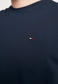 Granatowa koszulka z bawełny z okrągłym dekoltem, z małym haftowanym logo w czerwonym, białym i niebieskim kolorze po lewej stronie klatki piersiowej. Miękka w dotyku.