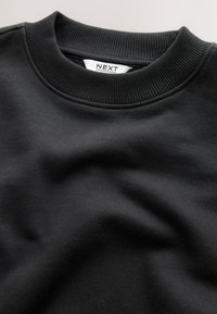Fekete pulóver bordázott, kerek nyakkivágással, sima textúrával és egy címkével, amely a "NEXT" márkanevet mutatja. Az anyag puhanak és tartósnak tűnik.