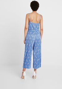 Modrá květinová kombinéza se širokými nohavicemi, nastavitelnými ramínky a přiléhavým pasem. Vyrobena z lehké tkaniny s hladkou texturou.