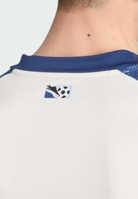 Weißes Sporthemd mit blauem Kragen und strukturierten blauen Akzenten auf den Schultern. Auf der Rückseite befindet sich ein Logo, das einen Fußballschuh und einen Ball zeigt.