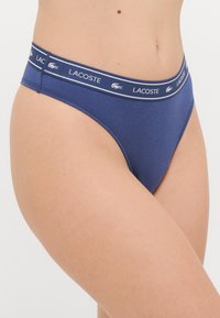 Tamsiai mėlynos trumpikės pagamintos iš minkštos medžiagos, su plačiu juosmeniu, kuriame matomas "LACOSTE" logotipas ir krokodilo emblema. Standartinio kirpimo dizainas.