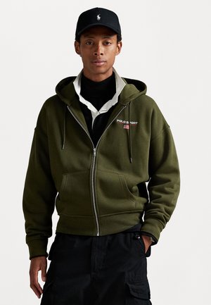 Oliven grøn zip-up hættejakke med forreste lommer og snøre i hætten. Sort cap med logo, lagdelt med hvid skjorte og sort rullekrave under.