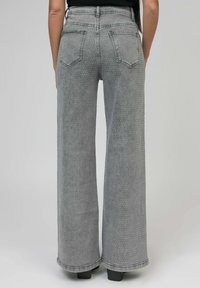 Personne portant un jean gris à taille haute et pattes d'éléphant avec des petits strass et des poches arrière, se tenant sur des chaussures à plateforme grises.