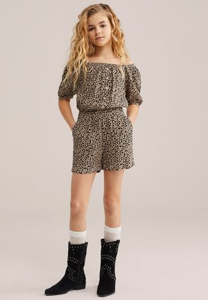 Ragazza giovane con lunghi capelli mossi che indossa una maglietta leopardata a spalle scoperte e pantaloncini, calzini bianchi e stivali neri borchiati, in piedi con le mani nelle tasche.