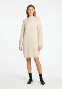 Beige stickad klänning med långa ärmar, hög krage och avslappnad passform. Försedd med en fransig fåll. Bärs med svarta strumpor och skor.