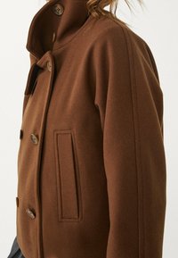 Brązowa kurtka polarowa z wysokim kołnierzem, podwójnymi guzikami, bocznymi kieszeniami i gładką fakturą. Proste, czyste linie.