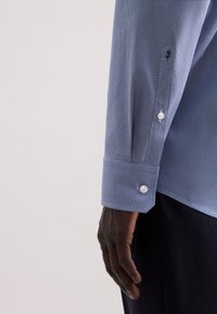 Camicia azzurro chiaro, con una texture e un motivo di piccole cerchi, dotata di bottoni bianchi e un design di polsino classico. Visibilmente ben adattata al polso.