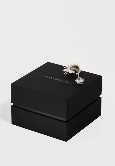 Srebrna uhan v obliki polmeseca z ostrimi konicami in sponko z jasnim dragim kamnom, prikazan na črni škatli za nakit z napisom ANNIEROETTI.