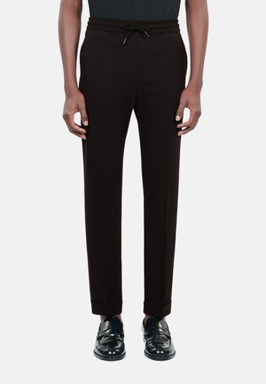 Czarne dopasowane spodnie z regulowanym paskiem na sznurku w talii, slim fit i nogawkami ze ściągaczami, wykonane z gładkiego materiału. Na nogach błyszczące czarne loafersy.