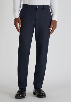 Marineblaue Baumwollhosen mit geradem Bein, Bund mit Knopfverschluss und Seitentaschen. Kombiniert mit schwarzen Slippern und einem weißen Langarmshirt.